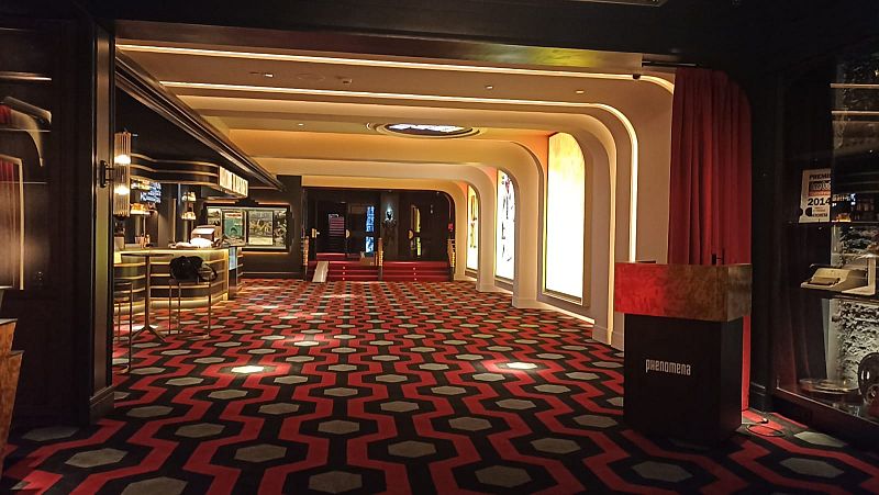 En el interior de un cine, se aprecia una barra con carteles y una vitrina con premios. El espacio se caracteriza por arcos iluminados, una alfombra con diseño y una iluminación cálida, sugiriendo un ambiente acogedor.