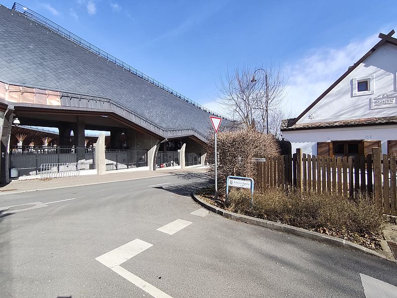 Una calle asfaltada en Felcsút, con una casa blanca de estilo tradicional húngaro y detalles en madera, junto a la entrada de un estadio. Se observa una señal de "Ceda el paso" y el año "2005" en la fachada de la casa.