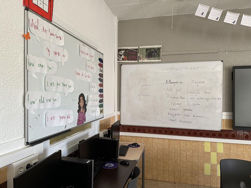 En un aula de idiomas, se observan dos pizarras blancas: una con preguntas y respuestas en inglés y un dibujo, y otra con palabras en catalán y su traducción al castellano. Varios ordenadores se sitúan sobre una mesa, y la pared está decorada con carteles.