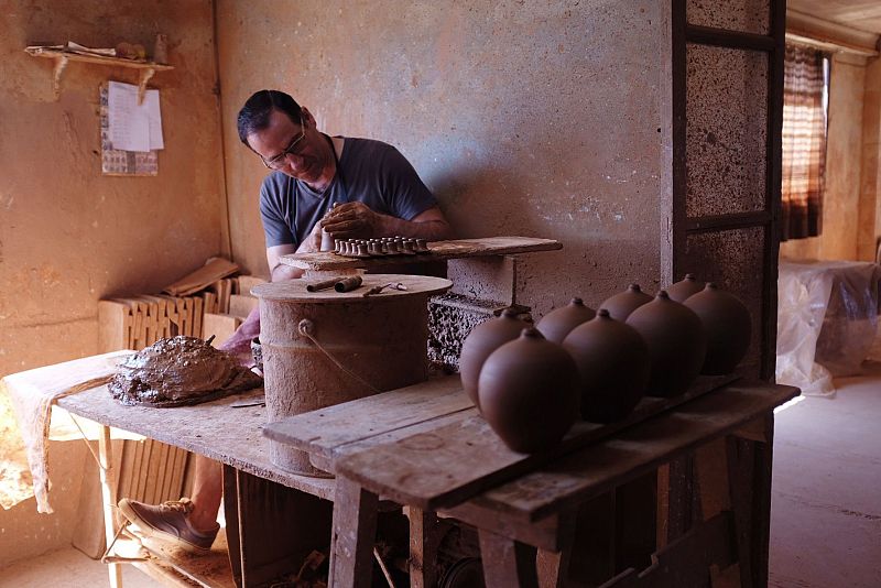 Alfarero en su taller modelando arcilla con herramientas y vasijas de cerámica sin esmaltar en un estante.