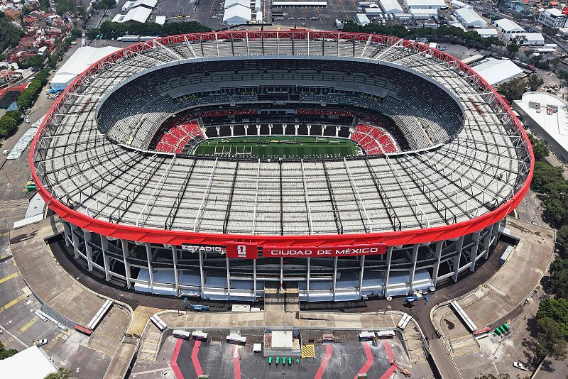 Estadio Azteca, Ciudad de México. Vista aérea cenital con techo de celosía, banda roja exterior y cartel "ESTADIO CIUDAD DE MÉXICO". Campo verde y gradas grises, blancas y rojas.