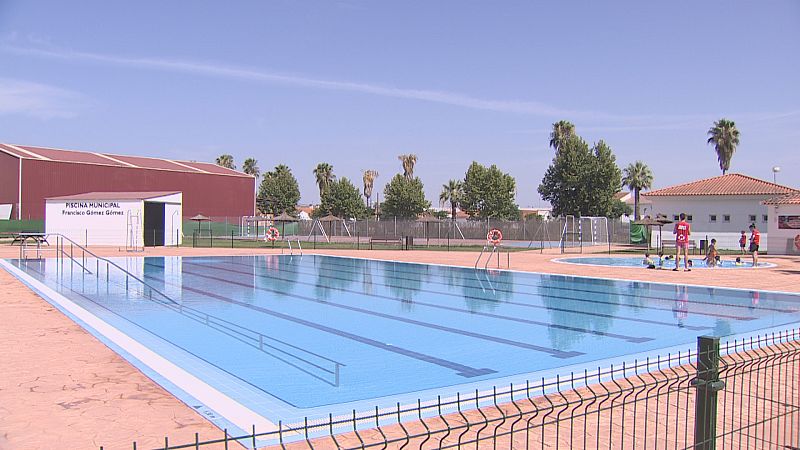 Vista panorámica de un complejo de piscinas municipales con dos piscinas, una rectangular y otra circular con niños. Se observan edificaciones, vegetación y mobiliario.