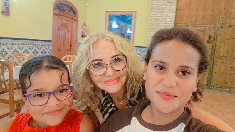Ana Garrido y sus dos niñas de acogida sonríen a la cámara en un interior con decoración tradicional, luciendo gafas y atuendos variados.