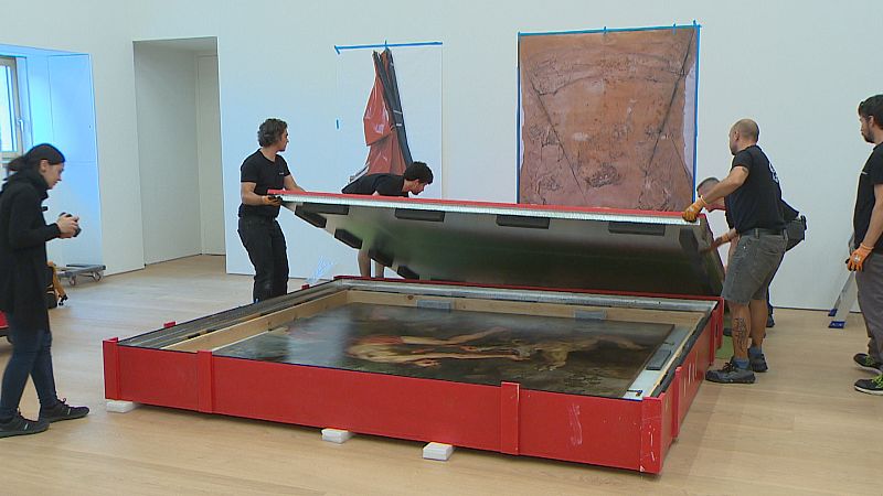 Un equipo desembala una pintura clásica de una caja roja en una galería. Una mujer documenta la escena con un dispositivo móvil mientras otros manipulan la tapa de la caja.