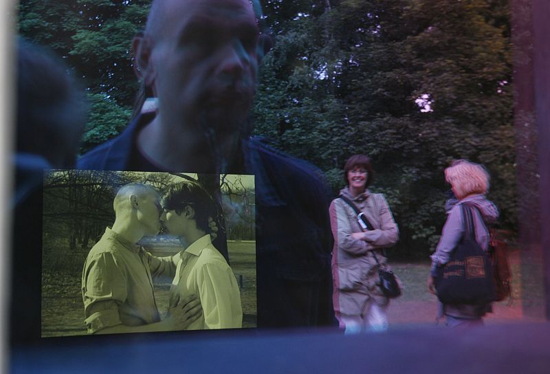 Gente reflejada en el cristal protector en el monumento en recuerdo de las víctimas gay del Holocausto junto a la pantalla de vídeo que muestra a una pareja homosexual besándose.