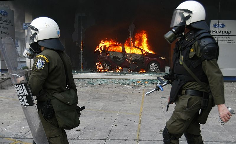 Policemen advance in front of a burning car inside a car dealership during riots in Athens