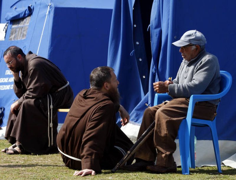 Un hombre habla con un monje en junto a una tienda de campaña del campo de L'Aquila.