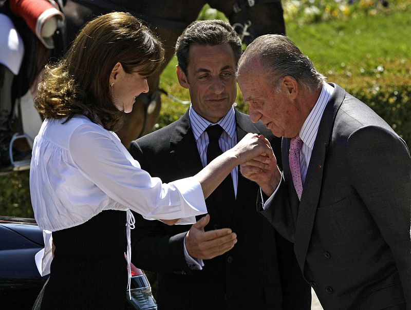 El Rey Juan Carlos I besa la mano a Carla Bruni delante de Sarkozy.