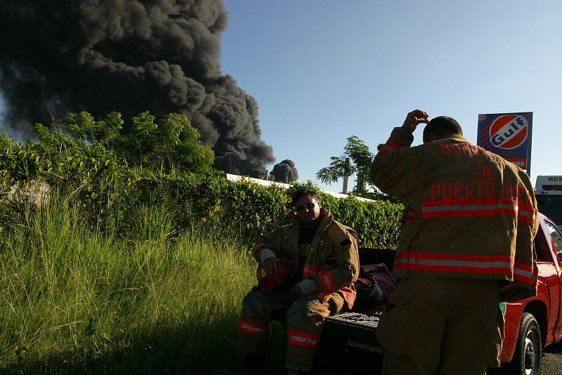 Bomberos descansan mientras se levanta una columna de humo debido al incendio de varios tanques de la refinería de petróleo de la empresa Caribbean Petroleum Corporation.