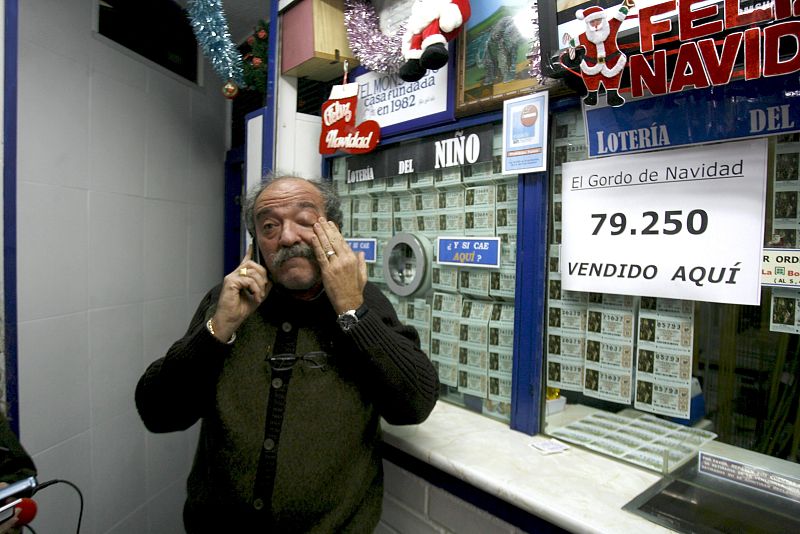 El propietario de la administración de lotería de la calle General Millán Astray de Madrid, con el cartel que anuncia que ha vendido el Gordo del sorteo extraordinario de Navidad, dotado con tres millones de euros a la serie.