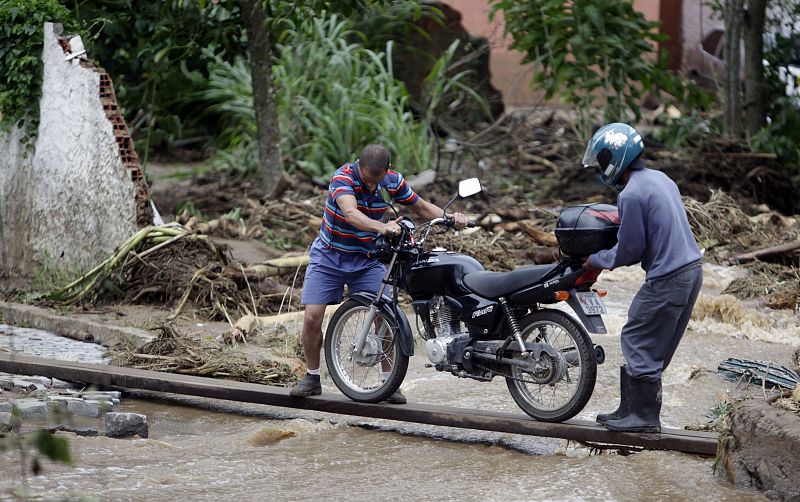 Dos hombres tratan de remolcar una moto a través de un trozo de madera sobre la carretera inundada.