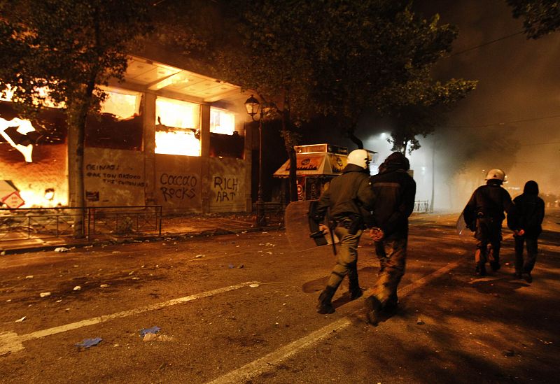 Agentes antidisturbios acompañan a dos detenidos mientras pasan por un edificio histórico de Atenas en llamas