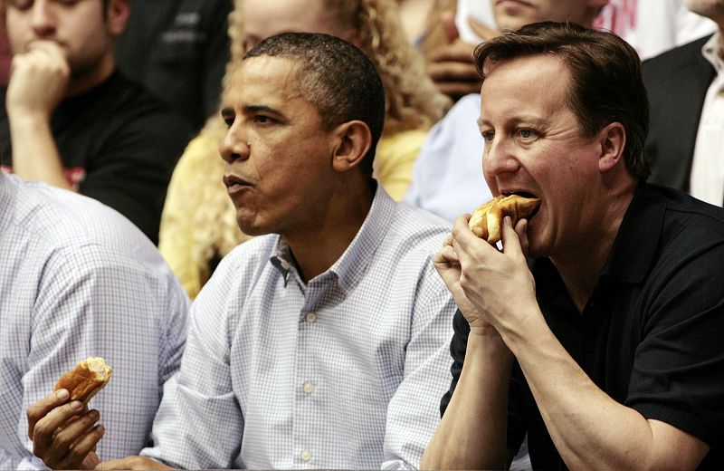 Obama y Cameron comen perritos calientes mientras disfrutan de un partido de baloncesto de la liga universitaria.