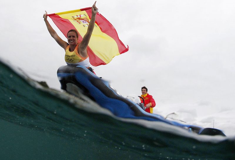 La española Marina Alabau celebra su victoria portando la bandera de España montada en su tabla de windsurf.