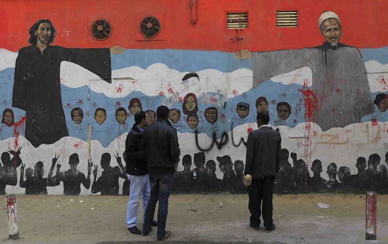 Viandantes miran un mural que representa las caras de manifestantes fallecidos. A la izquierda se representa a un clérigo copto, y a la derecha a uno musulmán.