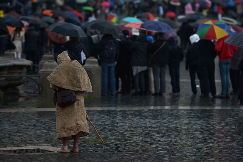 Un peregrino con los pies descalzos se mantiene solo bajo la lluvia, separado del grupo que observa el inicio del cónclave en la pantalla gigante