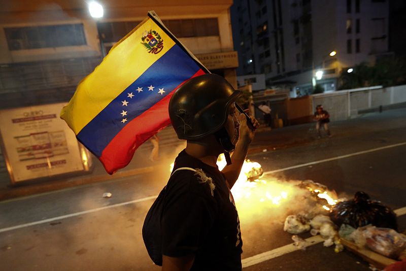 Un manifestante de la oposición camina levantando una bandera venezolana por delante de una barricada de fuego.