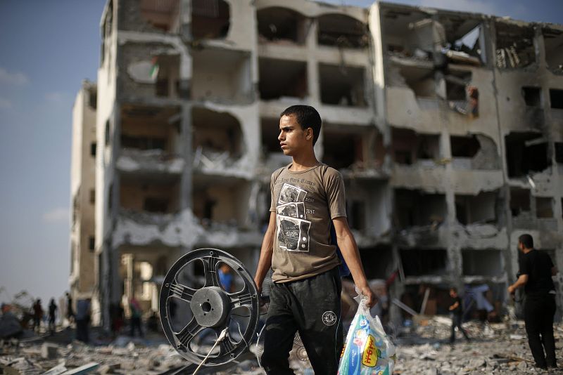 Un joven palestino carga sus pertenencias al pasar por un edificio muy dañado por los bombardeos en Beit Lahiya, al norte de la Franja de Gaza