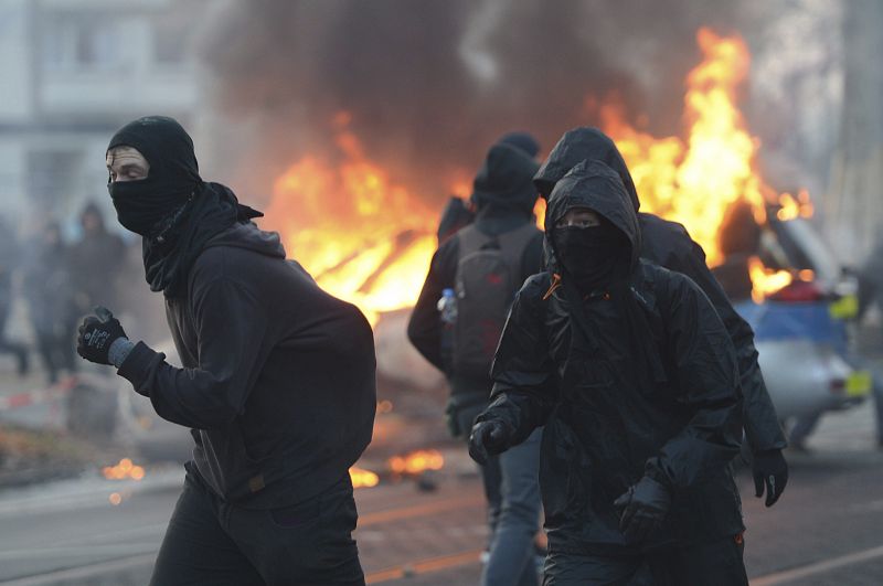 Manifestantes se cubren el rostro junto a un vehículo policial que arde