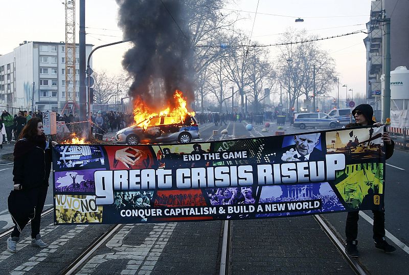 Dos manifestantes anticapitalistas portan una pancarta delante de una barricada