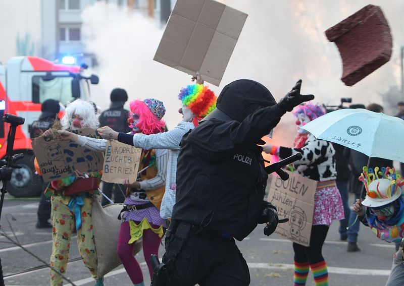 Un policia retira una piedra ante varios manifestantes vestidos de payasos