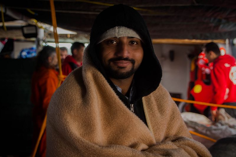 Un hombre posa con la manta que le acaban de facilitar los voluntarios de OPEN ARMS quienes trabajan al fondo