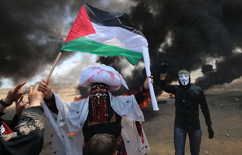 Unos manifestantes palestinos con máscaras ondean la bandera de Palestina entre neumáticos ardiendo