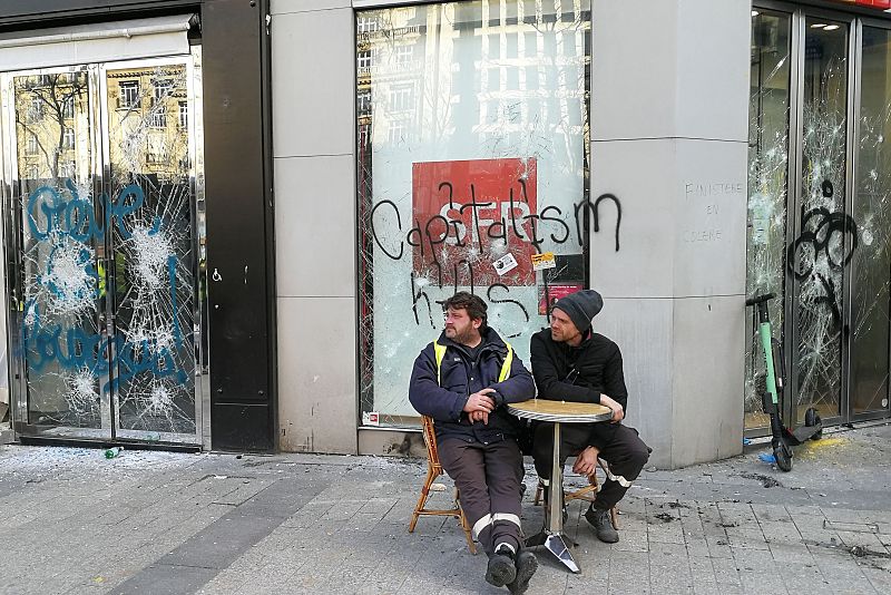 Dos manifestantes sentados frente a un escaparate dañado en los Campos Elíseos