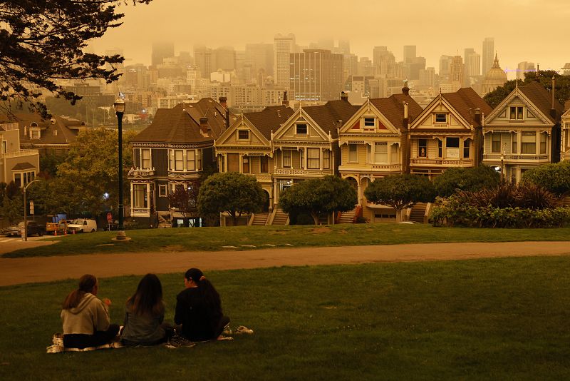 Varias personas se sientan en Alamo Square bajo un cielo nublado naranja y amarillo con vistas a The Painted Ladies, la icónica fila de casas victorianas históricas con un telón de fondo en el centro.