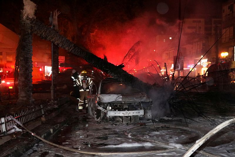 Bomberos israelíes caminan junto a un automóvil caído después de que fuera alcanzado por un cohete disparado desde Gaza hacia Holon, Israel.