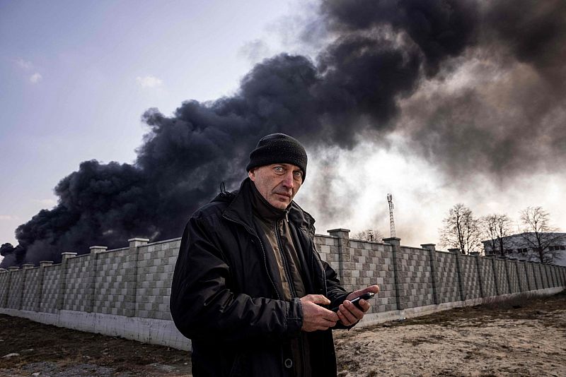 Un hombre mira su teléfono después de que los bombardeos hayan golpeado una planta de almacenamiento de combustible en Kalinovka, alzando una enorme columna de humo.