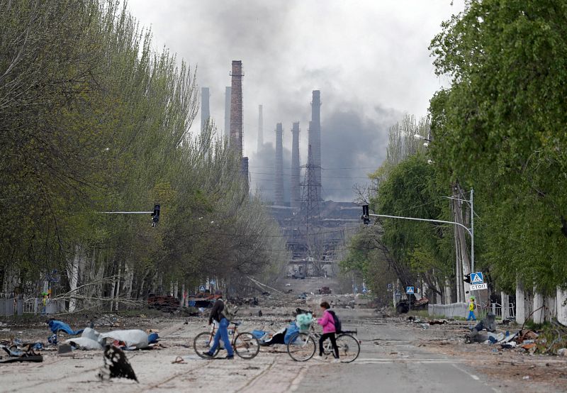 Un grupo de personas cruza la calle con sus bicicletas mientras el humo se eleva por encima de una planta de Azovstal durante el conflicto entre Ucrania y Rusia en la ciudad portuaria de Mariúpol, al sur del país.