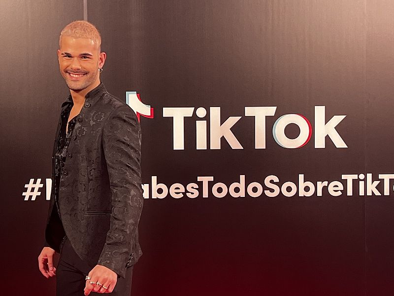 Enrique sonríe a la cámara y lleva un traje de chaqueta negro y el pelo decolorado. Posa delante de un cartel de TikTok.