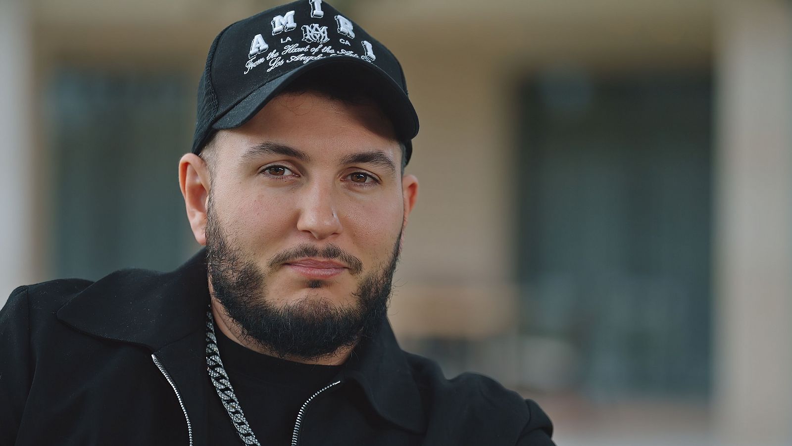 Omar Montes vestido de negro, y con una gorra en la cabeza. Sale en primer plano sonriendo.