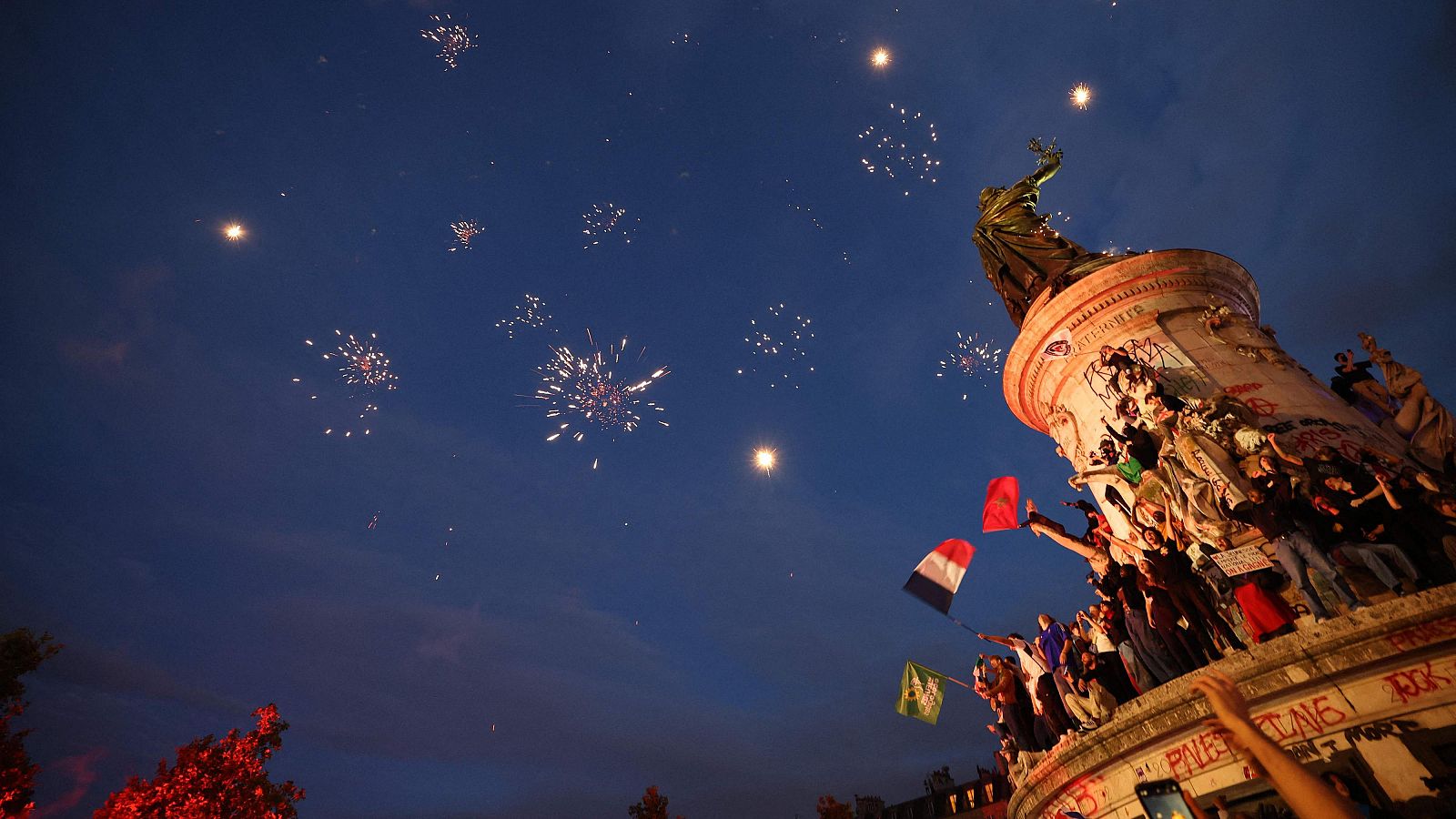 Varias personas ondean banderas, entre ellas la de Francia, mientras los fuegos artificiales iluminan el cielo