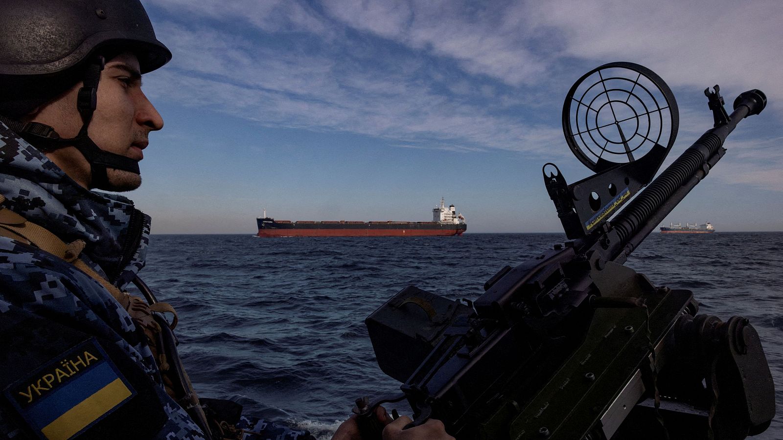 Un militar de la guardia costera de Ucrania maneja un arma en un barco patrullero mientras un buque de carga navega por el Mar Negro