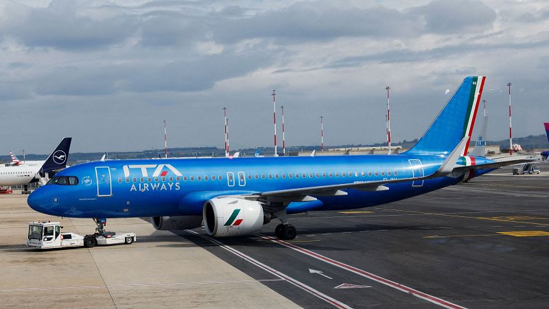 Avión de ITA Airways en pista de aeropuerto, remolcado por vehículo de tierra, con otro avión al fondo.