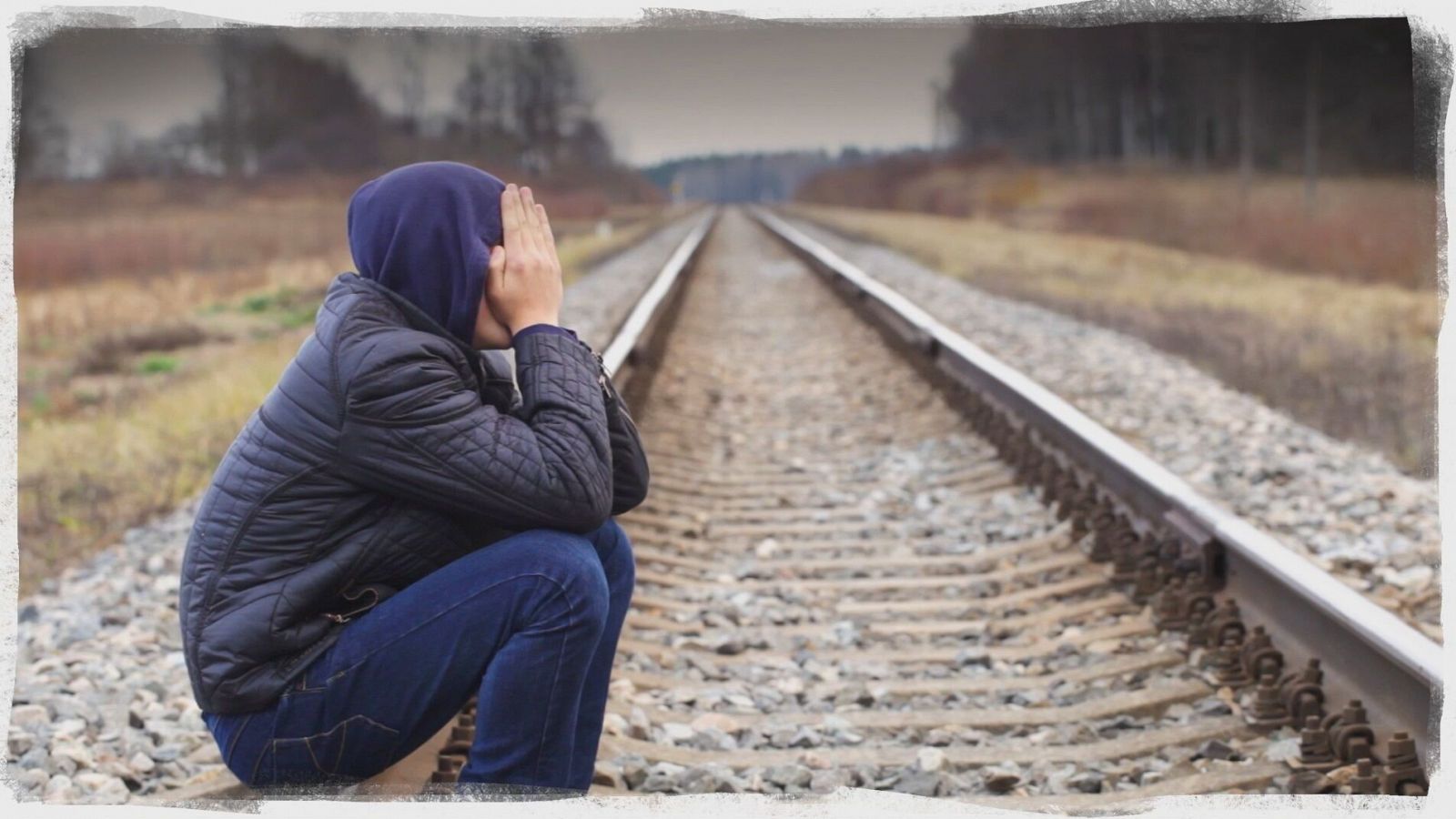 Adolescente con capucha y vaqueros se cubre el rostro junto a una vía de tren, mostrando tristeza y desesperanza.