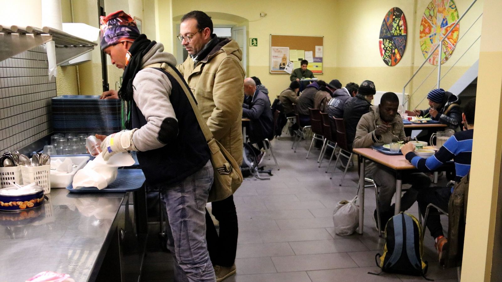 Comedor social La Sopa en Barcelona durante la cena, con varias personas sentadas y otras recogiendo sus platos.