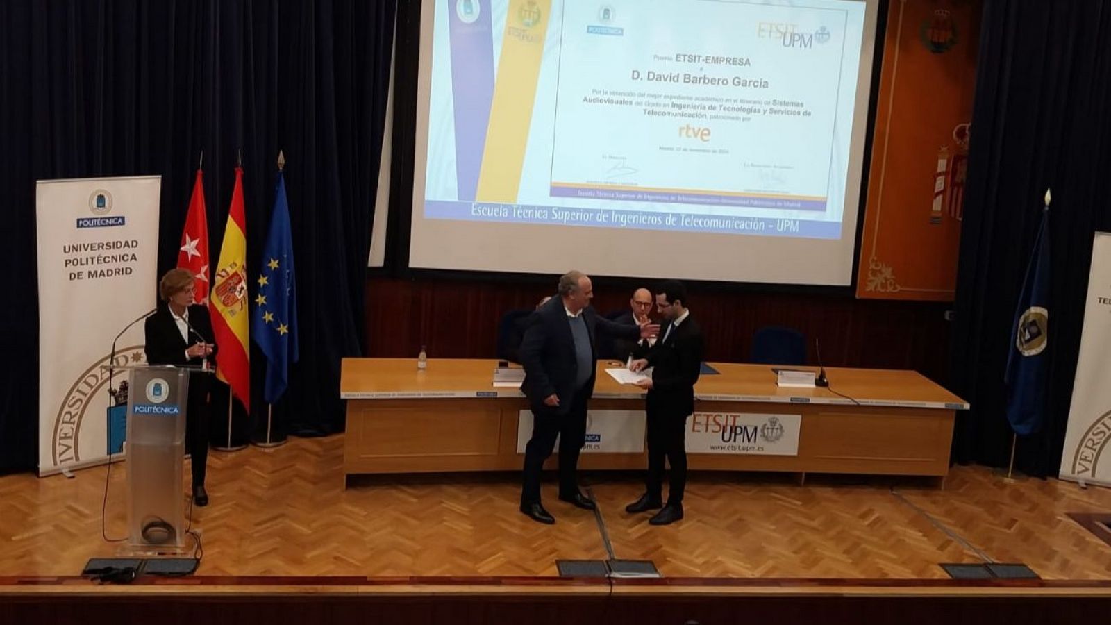 Entrega de premio Cátedras RTVE a David Barbero García en la ETSIT-UPM. Esteban Mayoral entrega el premio; presentes banderas de España, UE y Comunidad de Madrid.