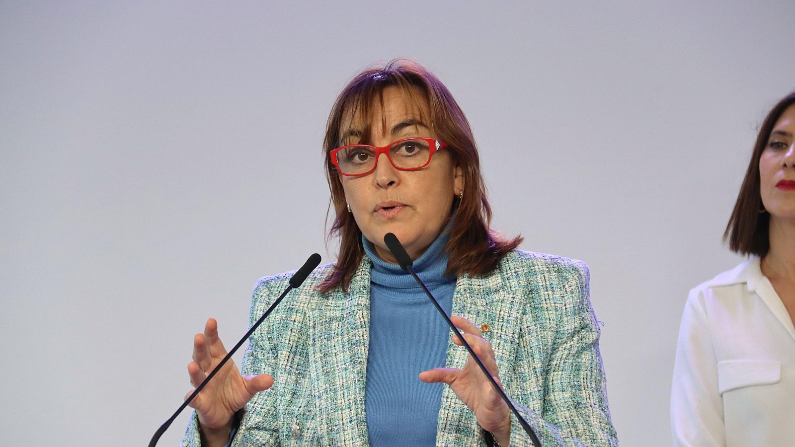 Silvia Paneque, consellera, habla en un evento. Lleva gafas rojas y chaqueta verde. Dos micrófonos están delante de ella.