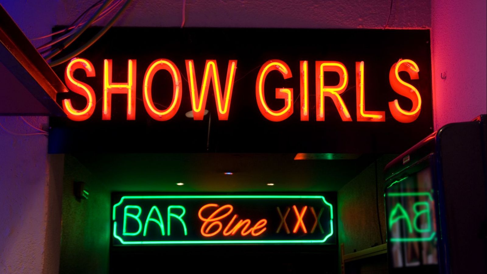 Letreros luminosos anuncian "SHOW GIRLS" y "BAR Cine XXX".  Reflejo de otro anuncio y cables eléctricos visibles.