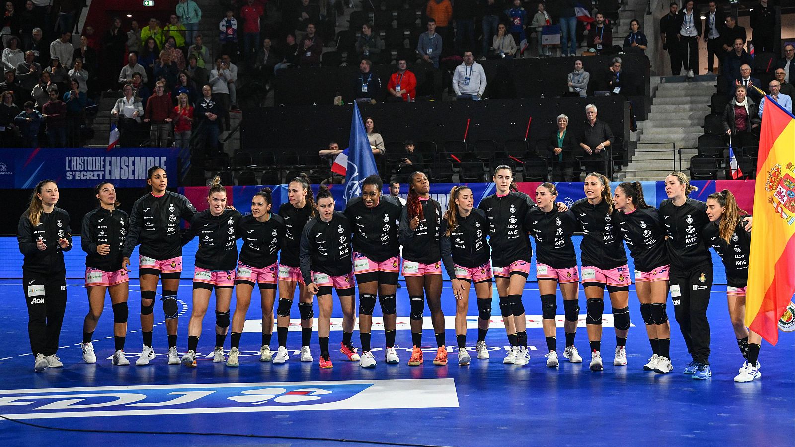 Selección española femenina de balonmano en formación, vestidas con sudaderas negras y pantalones cortos rosas. Una jugadora porta la bandera de España.