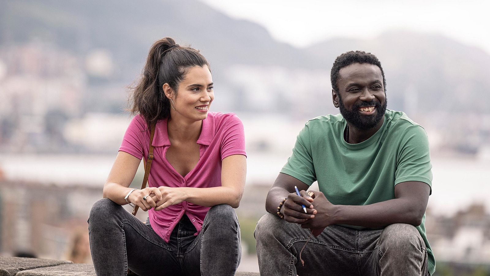 Dos personas sonrientes conversan en un muro.  Él lleva camiseta verde, ella rosa.  Parece una escena de 'Detective Touré'.