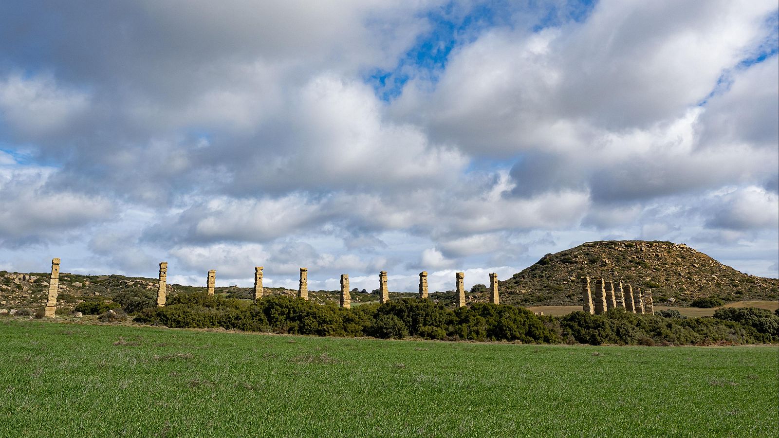 Ruinas de letrina romana en un campo verde, con columnas de piedra y una colina al fondo bajo un cielo nublado.