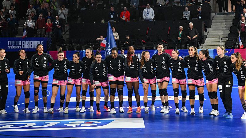 Las Guerreras, selección española femenina de balonmano, en formación. Visten sudaderas negras y pantalones rosas.  Se observa público y bandera francesa.