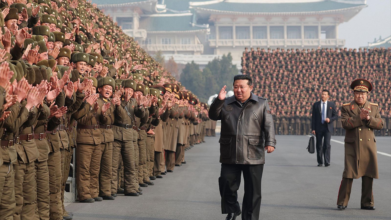 Kim Jong-un saluda a soldados norcoreanos en un desfile militar.  Un hombre en traje le sigue.  Edificio tradicional al fondo.