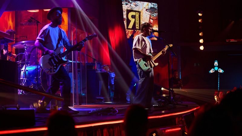 Actuación de Dani Fernández y su banda en La Revuelta de Broncano.  Dani toca la guitarra eléctrica en un escenario iluminado con luces rojas y azules.