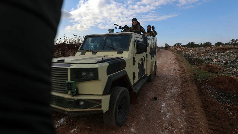 Vehículo militar camuflado transportando combatientes de Hayat Tahrir al-Sham en zona rural. Uno porta una ametralladora.