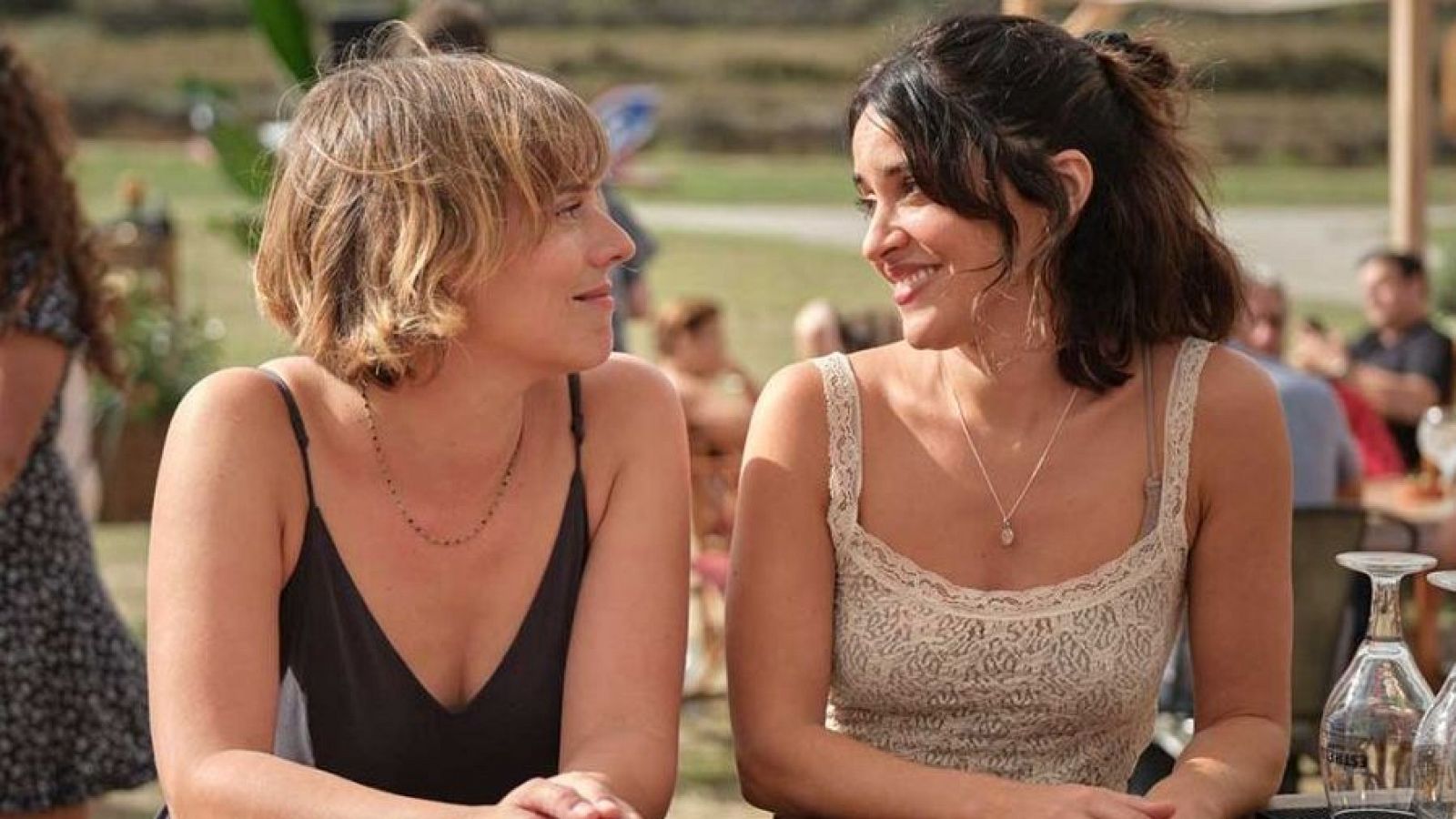 Dos mujeres conversan en una reunión al aire libre. Una lleva camiseta negra, la otra beige.  Ambiente festivo.